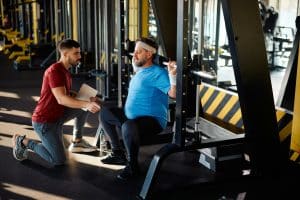 Mature man working out on exercise machine during exercise class with personal trainer in a gym.